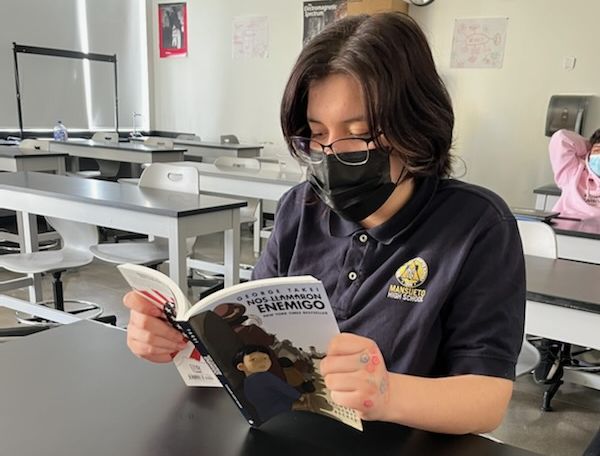 Photo shows Mansueto High School student reading George Takei's "They Called Us Enemy" in Spanish, book title reads "Nos Llamaron Enemigo"