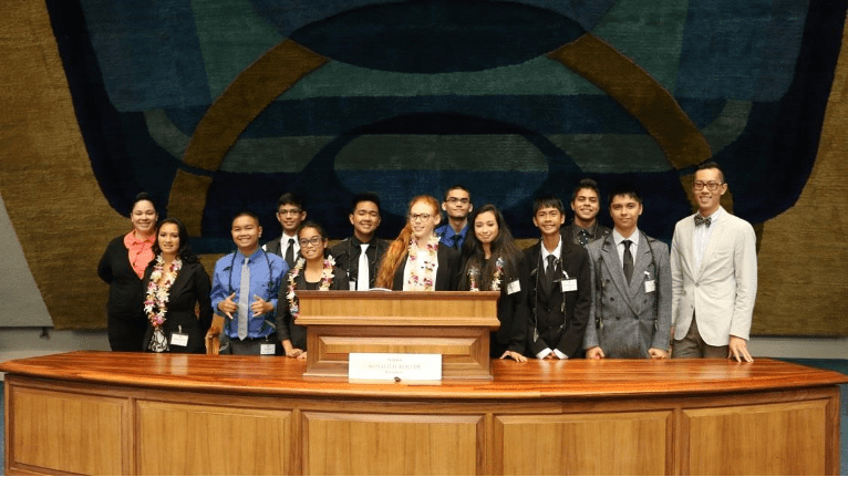 Photo shows JY Sun and his students in the Youth and Government program, standing behind a big official desk and podium in the Hawai'i state capitol in Honolulu