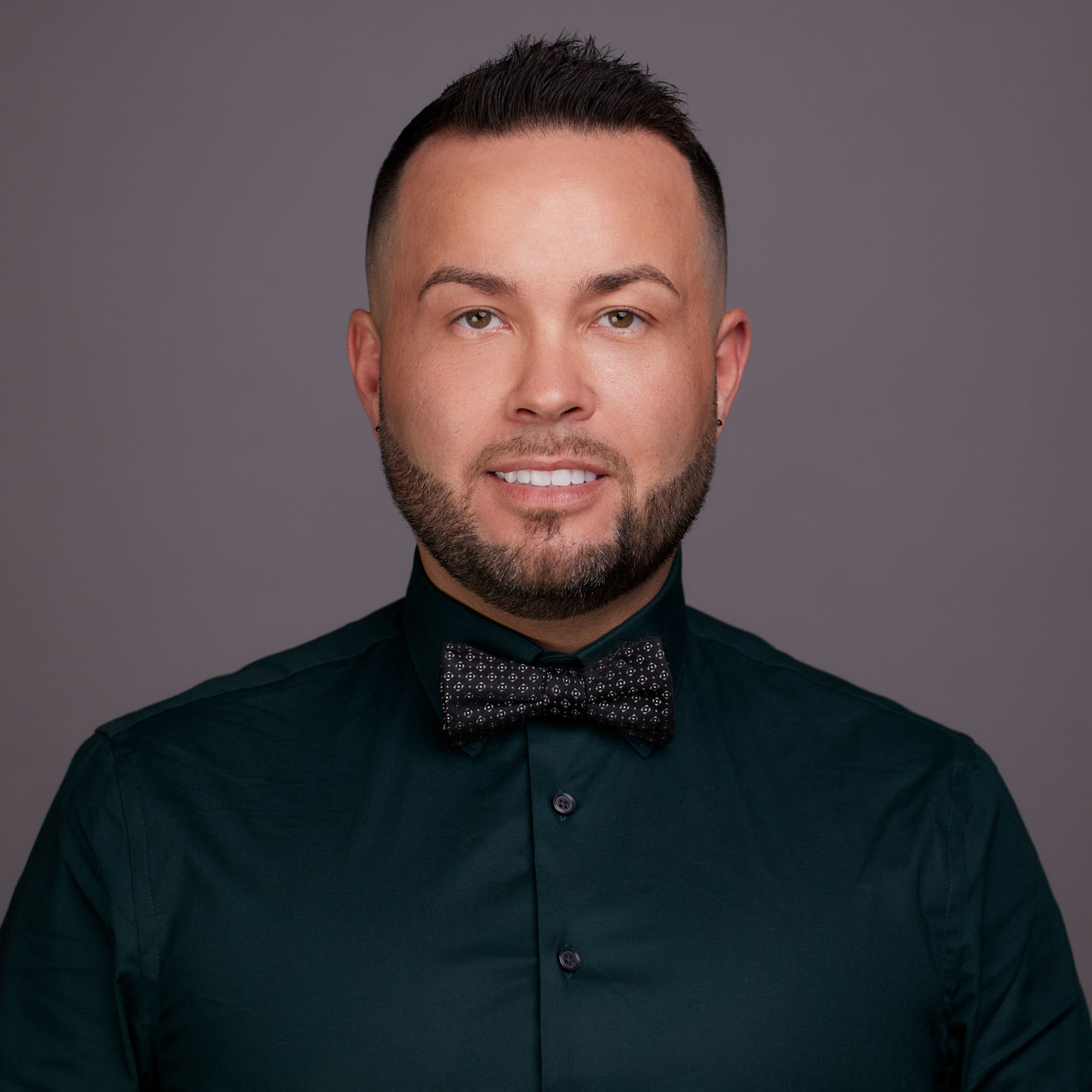 Image shows a headshot of Chase Johnson, new principal at Muchin College Prep. He is a young man with short brown hair and a short beard. He is wearing a long sleeved black button up. The background is a solid gray color.
