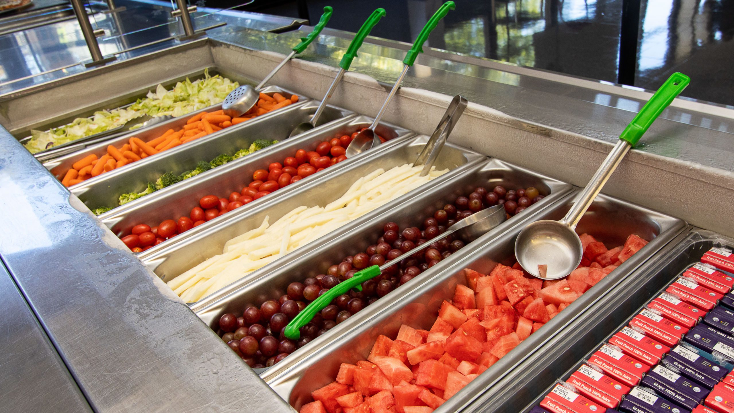 Photo shows a school cafeteria line at Noble Schools with fresh watermelon, grapes, carrots. broccoli, cherry tomatoes, lettuce, and jicama in the pans