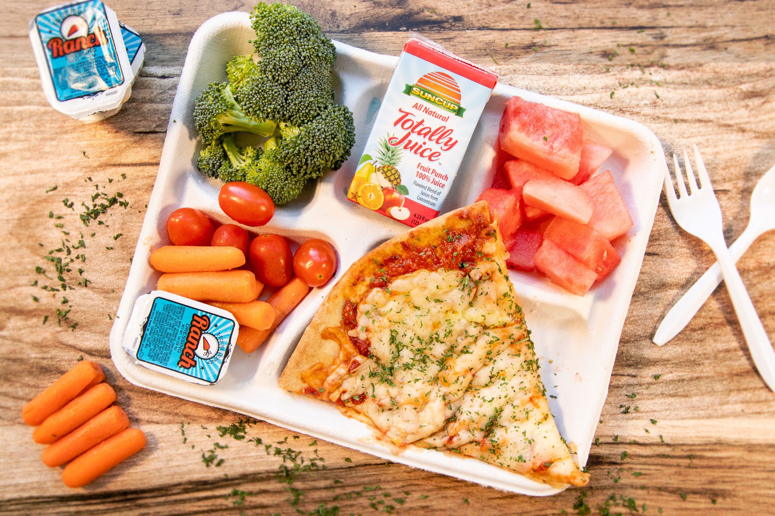 Photo shows a cafeteria tray with pizza, watermelon, broccoli, cherry tomatoes, and carrots on it. There is also utensils, more carrots, and ranch dressing to the sides of the tray.