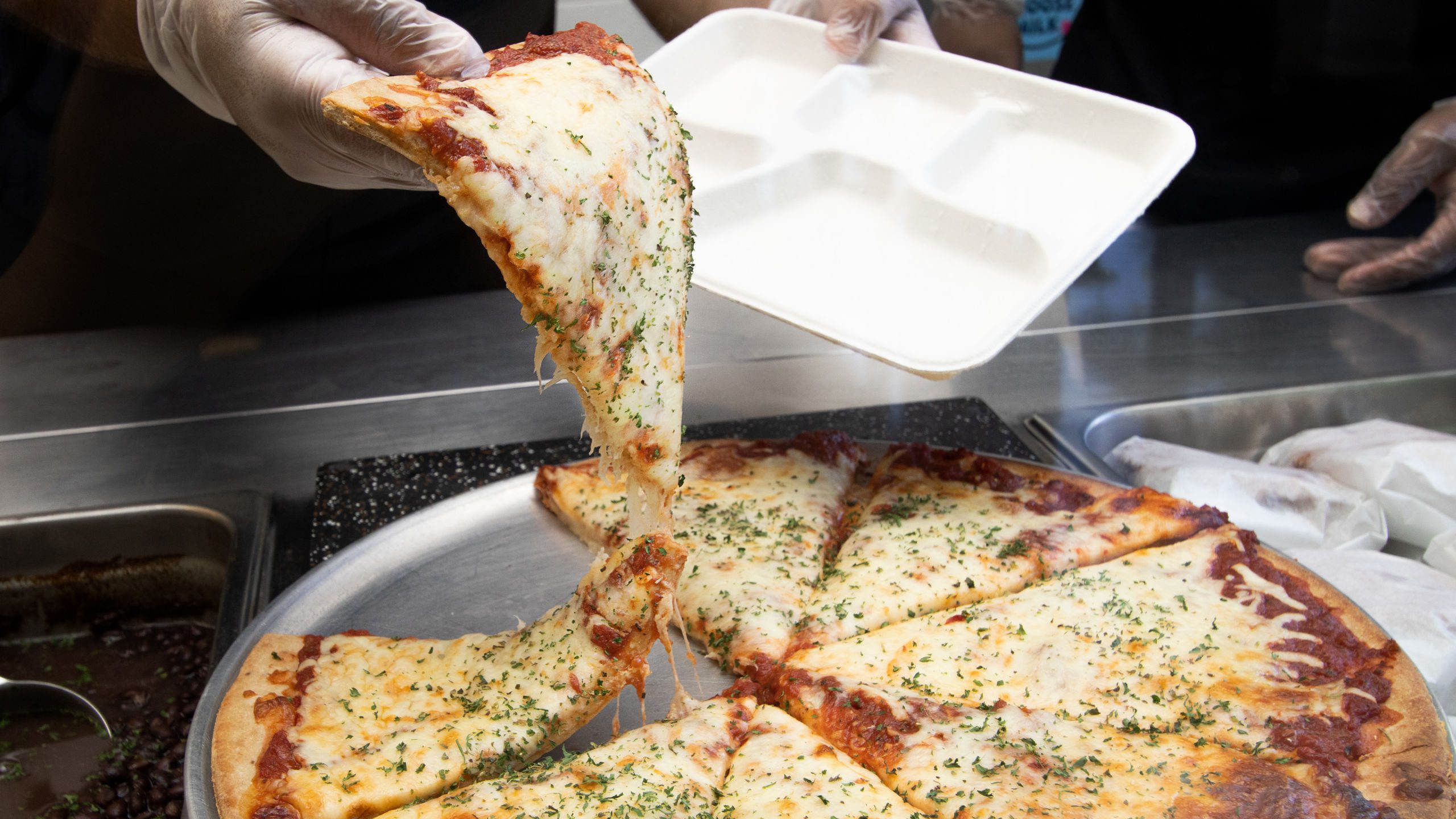 Photo shows a hand pulling a slice of cheese pizza off the pan and onto a compostable tray at Noble Schools