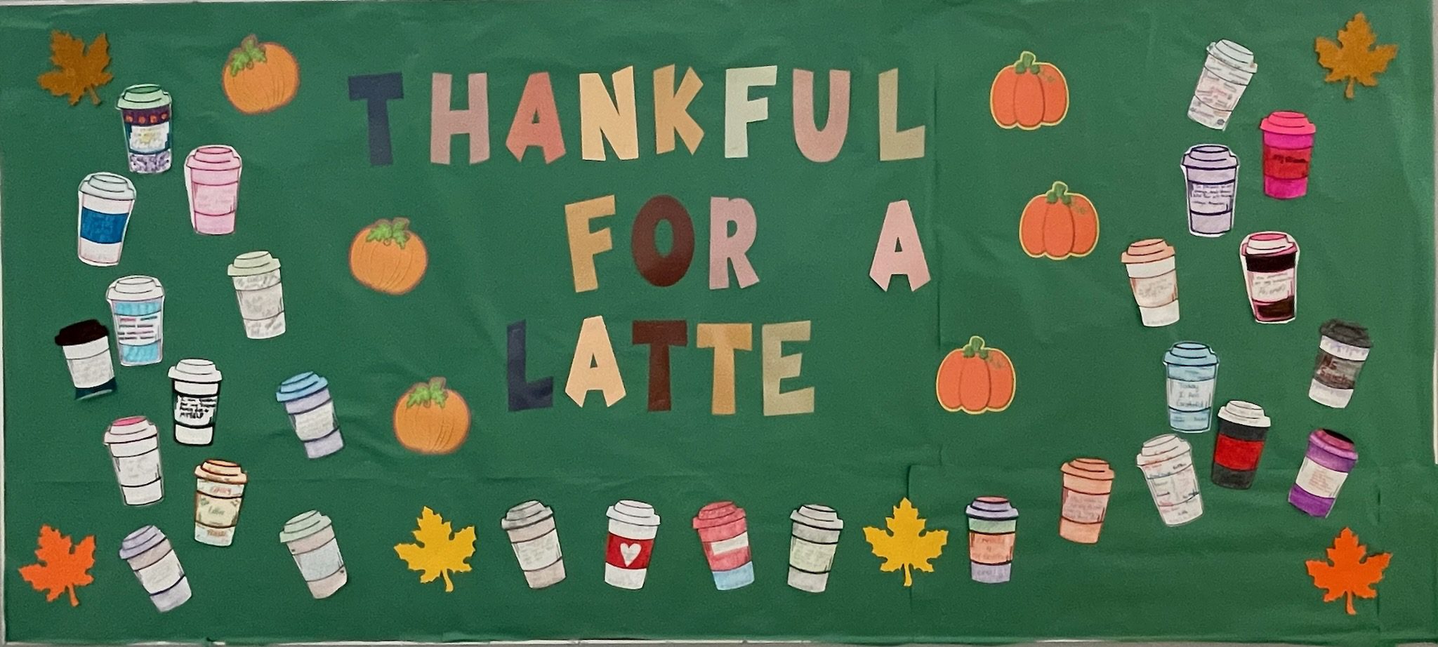 This image shows a dark green bulletin board that has the text "Thankful for a Latte" on it. The words are surrounded by paper drawings of pumpkins, leaves, and latte cups.