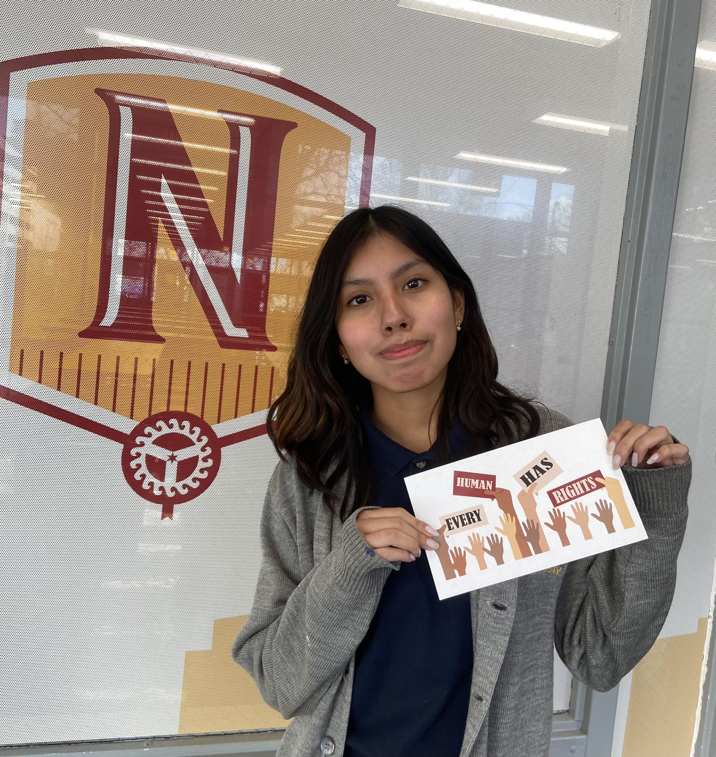 Image shows The Noble Academy student Leslie Caguana posing in front of a decal of The Noble Academy logo. She is holding a small white sign that has hands of all colors that are holding up mini signs that read "Every Human Has Rights"