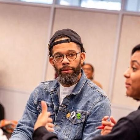 Photo shows Nicholas Jones participating in University of Chicago's Civic Actors Studio. He is wearing a jean jacket and a black snapback and listening to a person next to him talk.