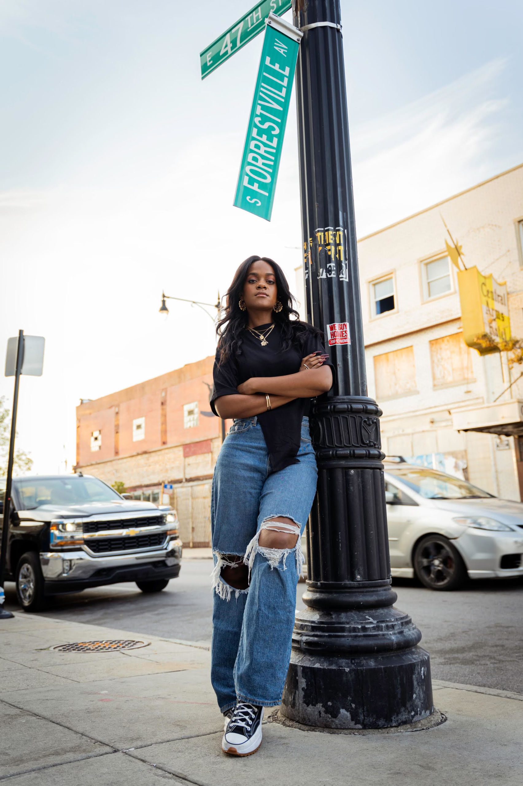 Photo shows a full body portrait of Michelle Thompkins, founder and CEO of The CornerStore Chicago. Michelle is a Black woman, wearing a black t-shirt, ripped jeans, and black Converse while leaning against a street lamp. Above her are street signs that read "South Forrestville Ave" and "East 47th Street". Behind her is a Chicago street with cars and buildings.