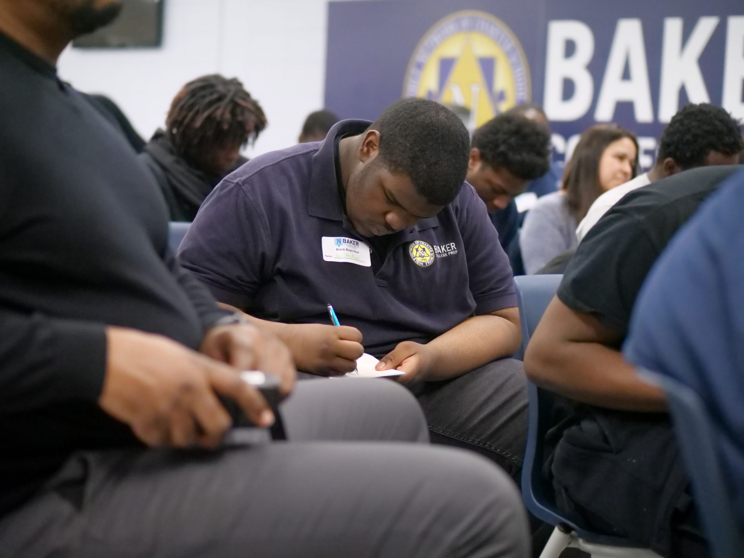 Image shows a young Black boy at Baker College Prep in Chicago, IL, writing in a journal during the Black Boys Heal: See Me, Hear Me event at his school.