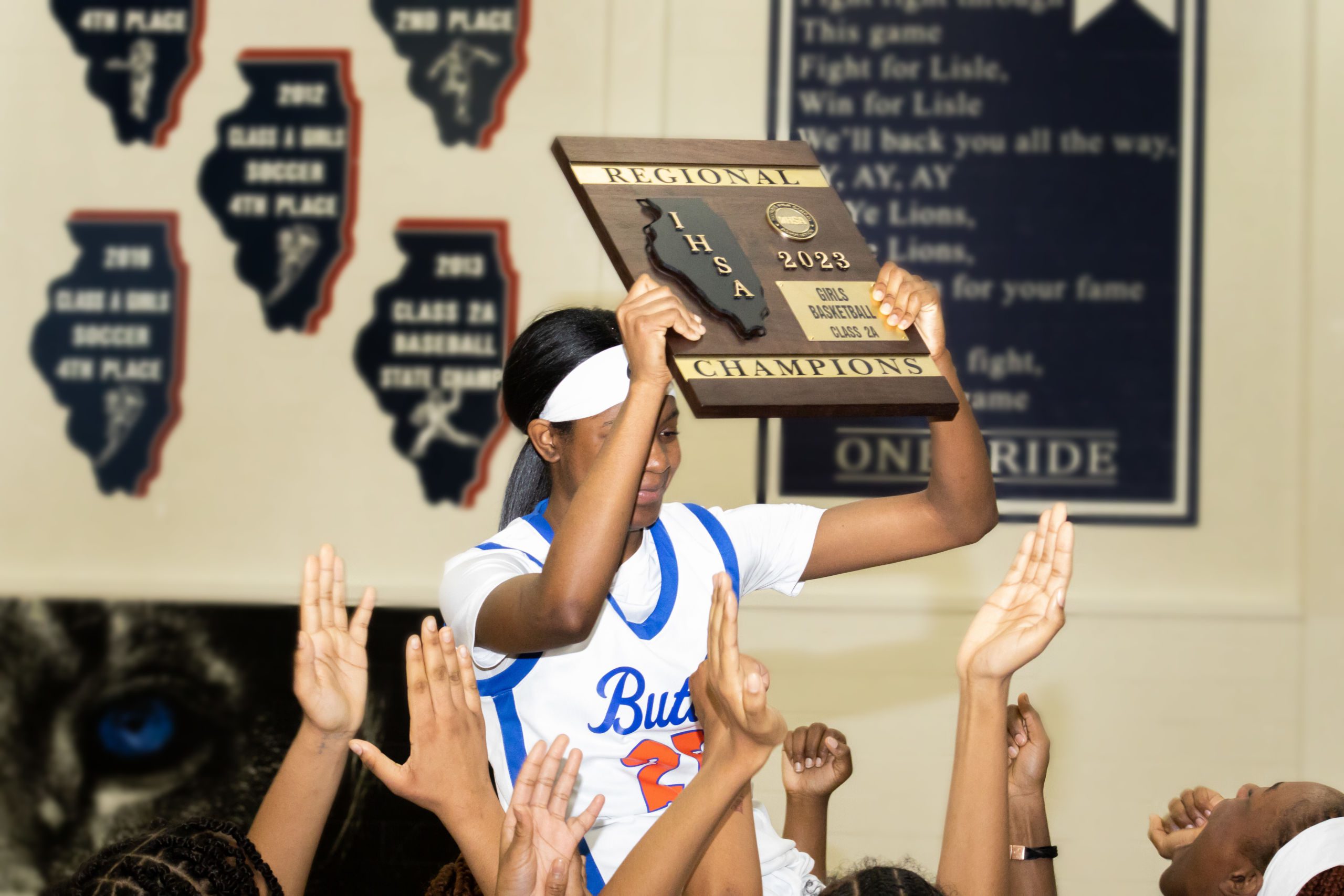 Image shows a basketball player on the Butler College Prep girls' basketball team, holding up the IHSA Regional Championship plaque while on the shoulders of her teammates after the Lady Lynx team won the championship for the second year in a row.