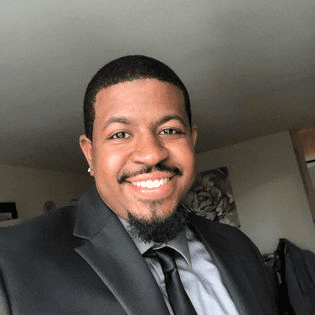 Photo is a headshot of Prentyce Robinson I, a teacher at Rauner College Prep. He is a Black man and is wearing a black blazer, gray collared shirt, and a black tie.