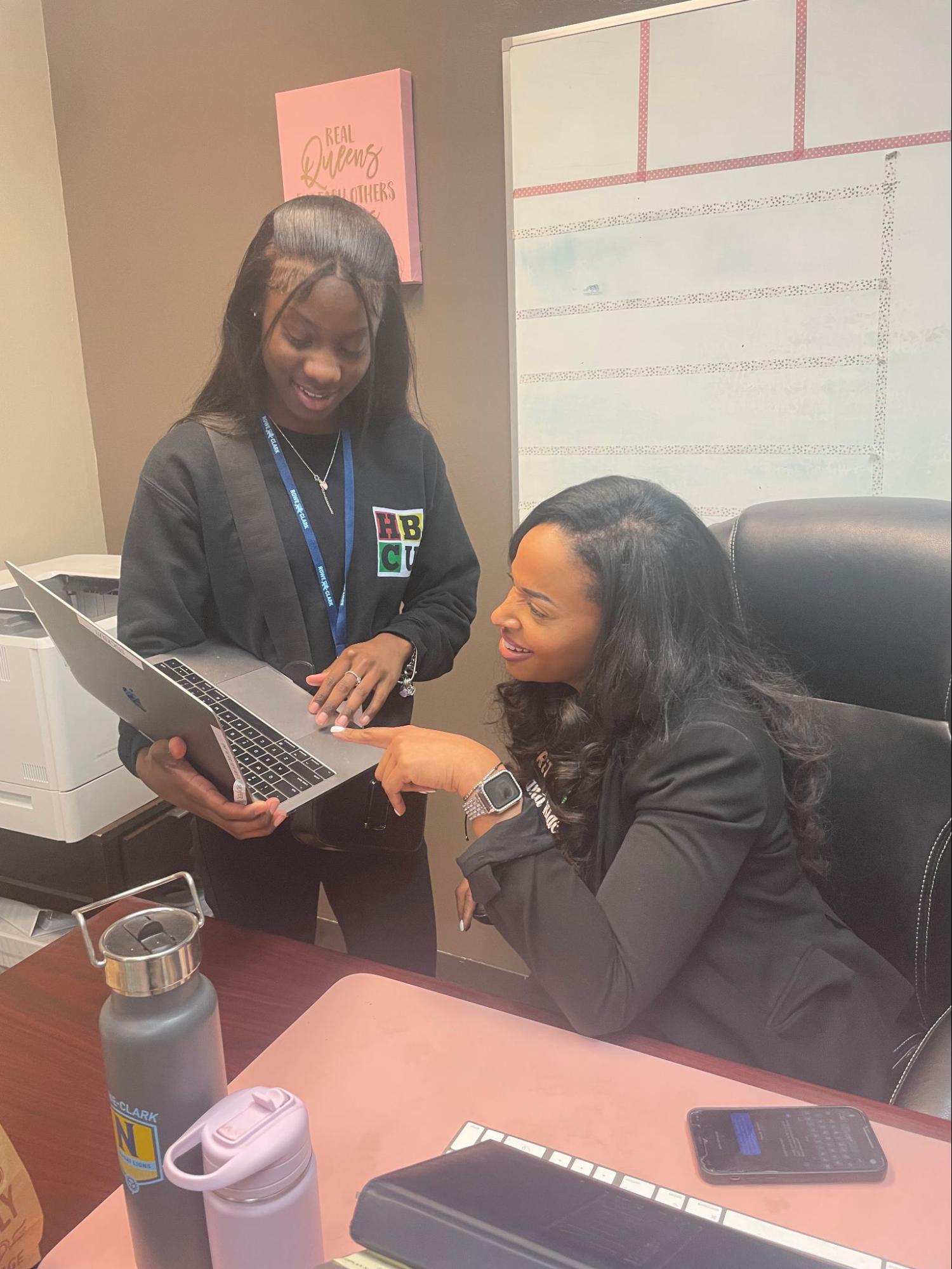 In this photo, Makya, a student, is standing behind a desk and showing what is on her laptop screen to her principal, Miracle Moss. Principal Moss is a Black woman with long, wavy black hair who is wearing a black blazer and is sitting in a black office chair. She is pointing at Makya's laptop as Makya stands beside her at her desk, holding her laptop.