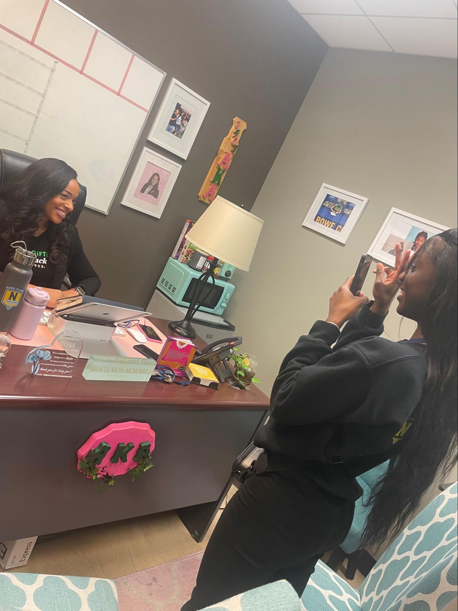 Makya stands in front of the principal's desk with a phone out. She is taking a picture of Principal Moss sitting and smiling at her desk for a social media post.