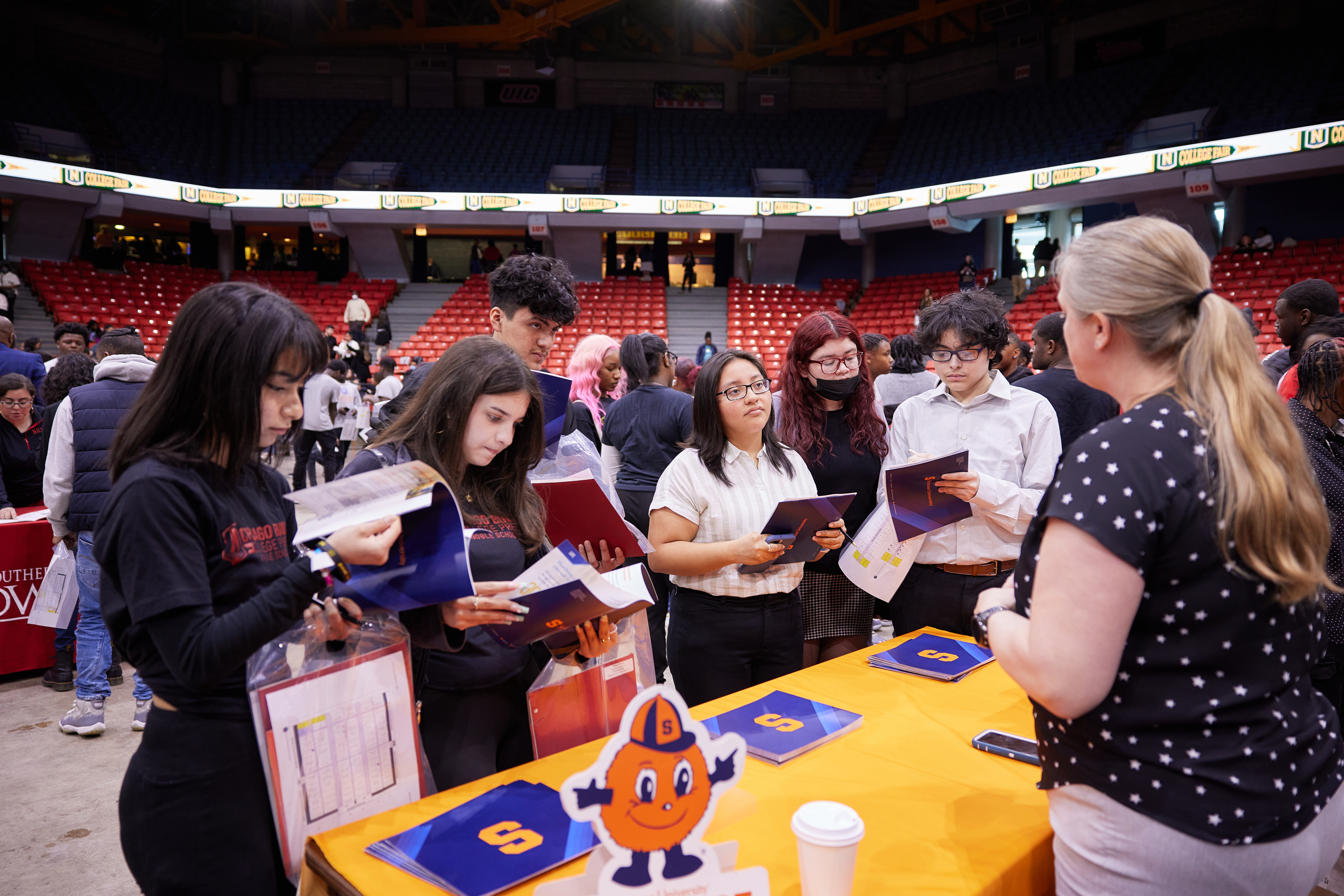 Students crowd around a college booth as they take notes, ask questions, and look at their pamphlet. The college recruiter has blonde hair in a pony tail and is facing towards the students, her face is not visible. Students are wearing their Noble Schools sweater that is a dark navy blue. The college booth table is yellow and has pamphlets and other college documents located.