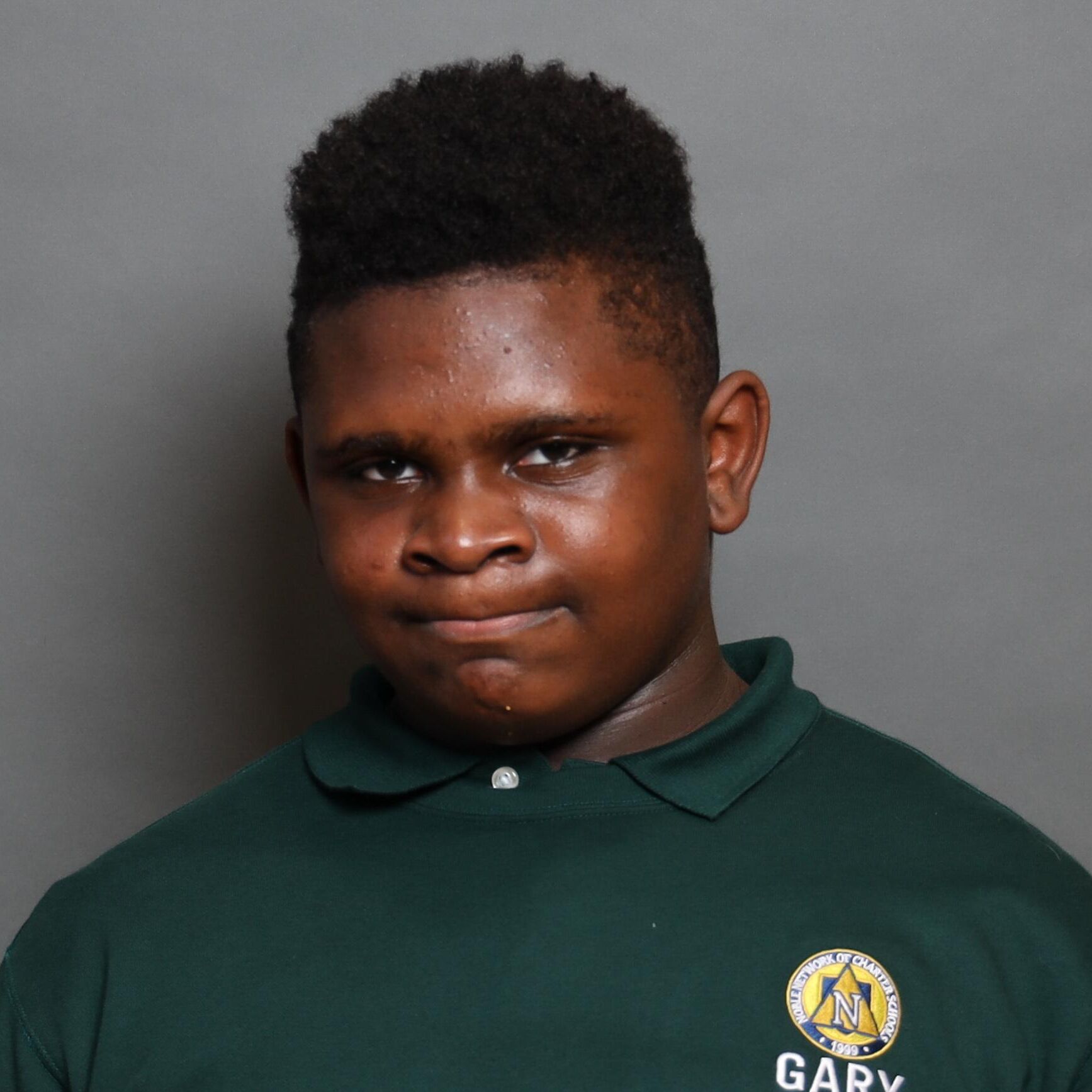 This photo shows a headshot of Dawson Harris, an 8th grader at Gary Comer Middle School in Chicago, IL. Dawson is a young Black boy with short dark brown hair, cut in a hi-top fade hairstyle. He is wearing a dark green shirt with the Gary Comer Middle School logo on it.