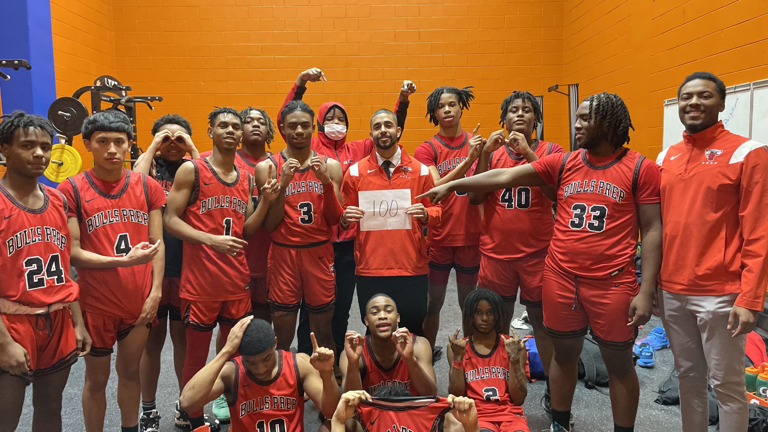 Chicago Bulls College Prep basketball students pose with Mr. Goldestein as they wear their red basketball gear. The students point to the sign Mr. Goldestein is holding that says "100" indicating their 100th win.