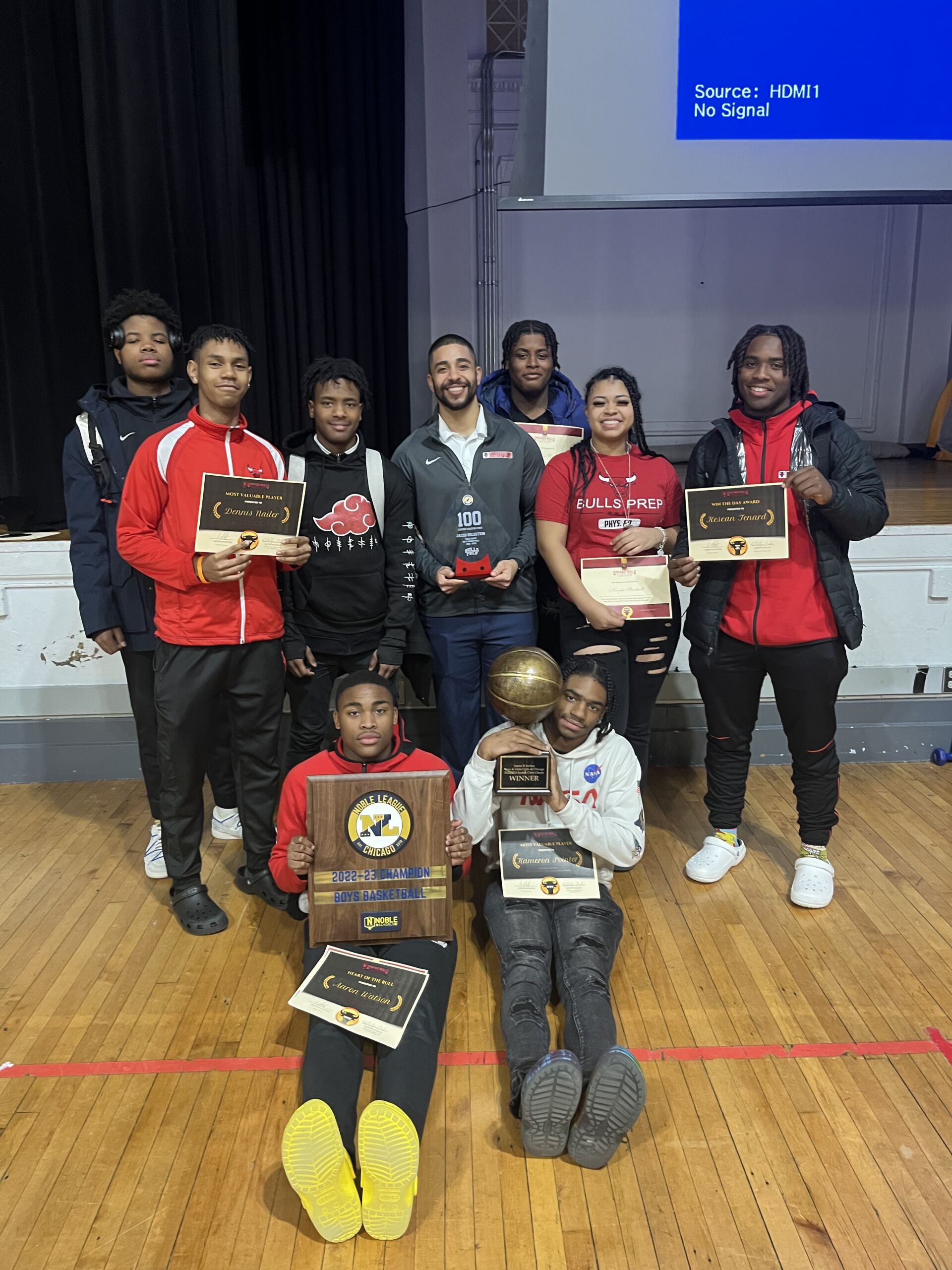 A few members of the Chicago Bulls College Prep basketball team pose with their trophies. They smile at the camera as they crowd around Mr. Goldestein, who is also holding an award for their 100th win.