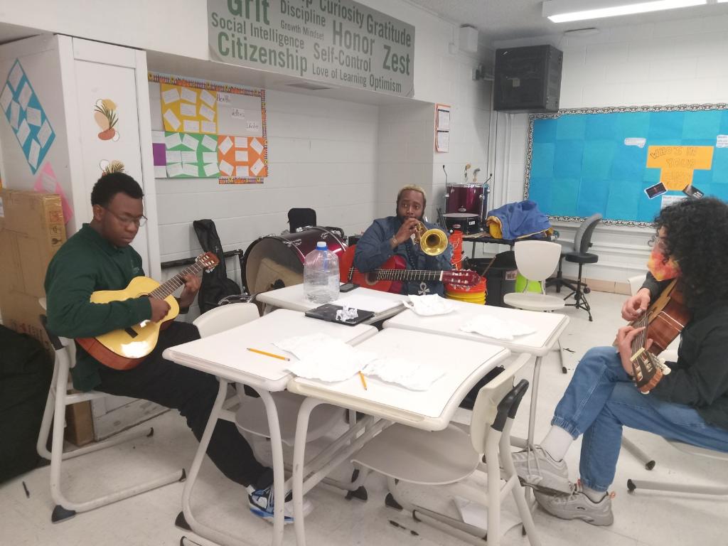 Image shows a scene of a classroom at Gary Comer Middle School. There is a grouping of four desks in the center and two students and one mentor sit around it with instruments. Tristan, a young Black boy with short dark brown hair and glasses, sit of the leftmost side, holding a guitar. To his right and in the middle is his mentor, Bobby, a Black man with a beard and short blonde hair. Bobby is holding a trumpet and has a guitar in his lap and is wearing a dark gray suit. To the right of him is another student holding a guitar.
