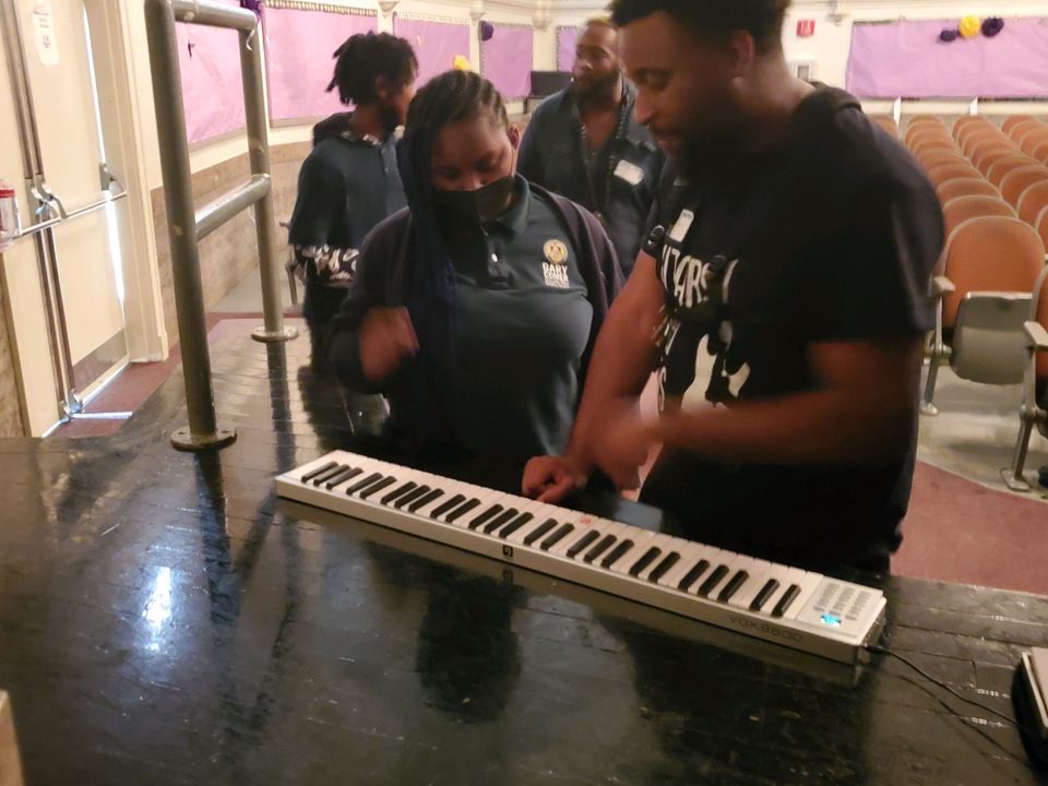 Photo shows two people standing over a keyboard. On the left is Mariah, a student at Gary Comer Middle School. Mariah is a young Black girl with medium-length dark brown braids. She is wearing a Gary Comer Middle School polo. On the right is Kevin, one of her music mentors from Guitars Over Guns. Kevin is a young Black man who is wearing a Black t-shirt with a white logo on it for Guitars Over Guns.