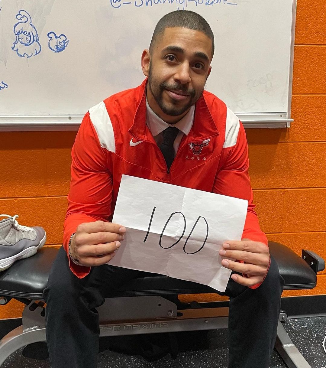 Jacob Goldstein poses for the photo. He is wearing a red & white, Chicago Bulls windbreaker jacket. He is holding a piece of paper with the number 100 written on it.