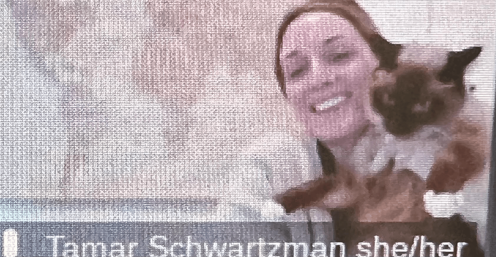 Ms. Shwartzman poses on Zoom with her cat. She has her hair tied back, wearing a cardigan sweater. She holds her Siamese colored cat next to her. The photo is a screenshot off of Zoom. The quality is slightly blurred but viewable.