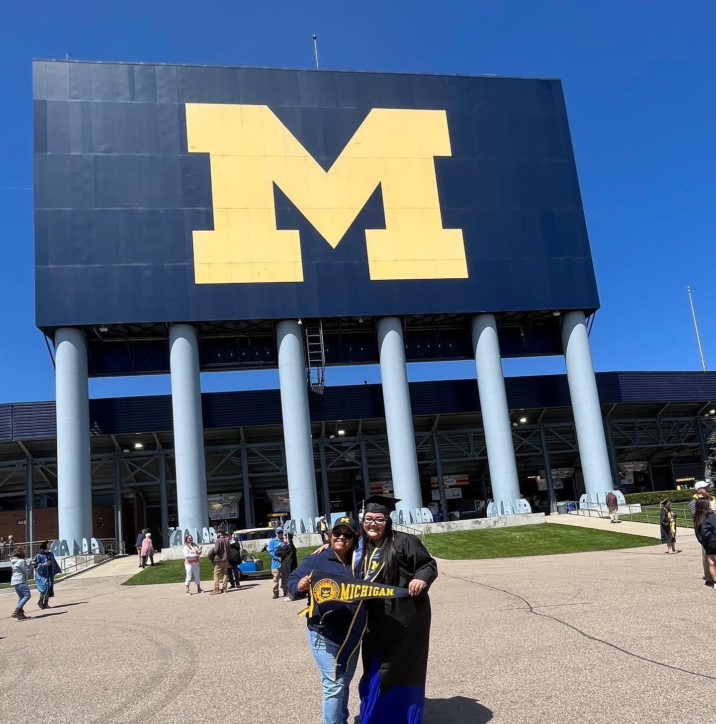 Vanessa Olivares, on the right, pictured with her mother, on the left. The two pose outside of University of Michigan at Vanessa's graduation. Her mother is wearing a University of Michigan hat, sunglasses, and blue jeans. Vanessa wears her graduation grown as both her and her mother hold University of Michigan banner flag.