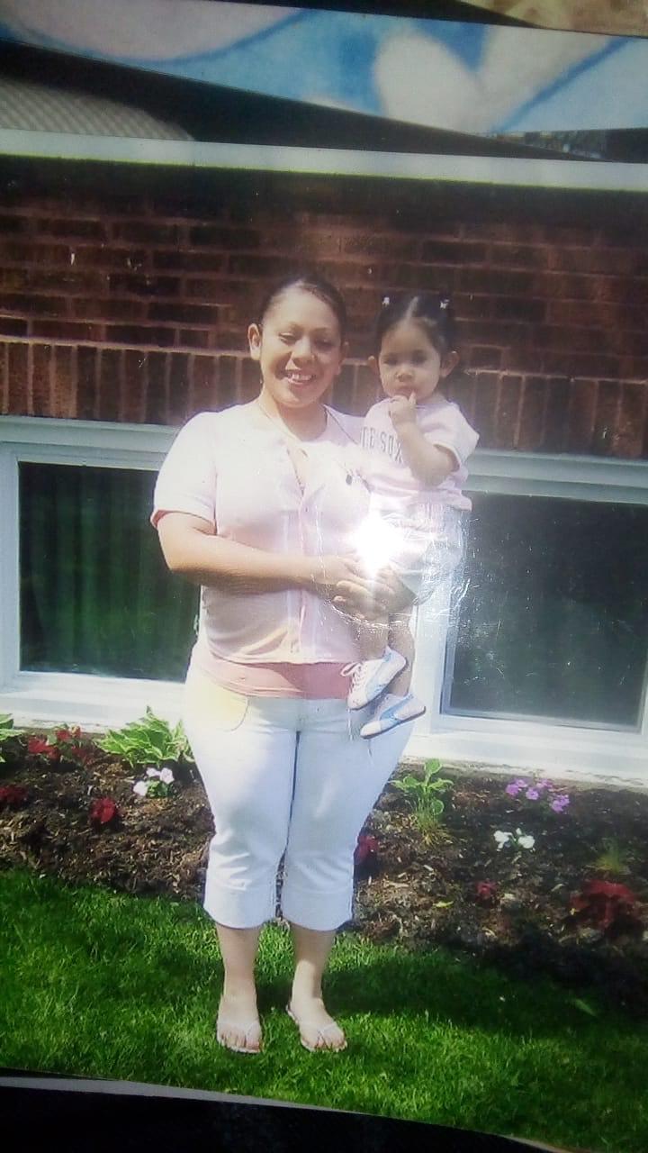 Harumi Palomino being held by her mother. Harumi is a baby and is wearing a pink white sox shirt. Her mother is wearing a pink baseball jersey and light blue jeans. Her hair is tied back as Harumi's hair is tied into multiple pig tails.