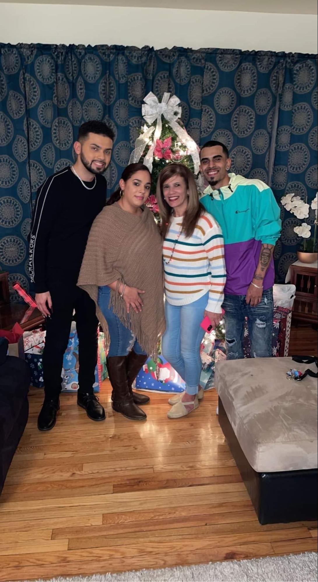 Cindy Sanchez with her family. She is wearing a brown cardigan and is posed next to her family members and her mother. Her mother is wearing a rainbow stripped shirt. The family poses in front of a Christmas tree.