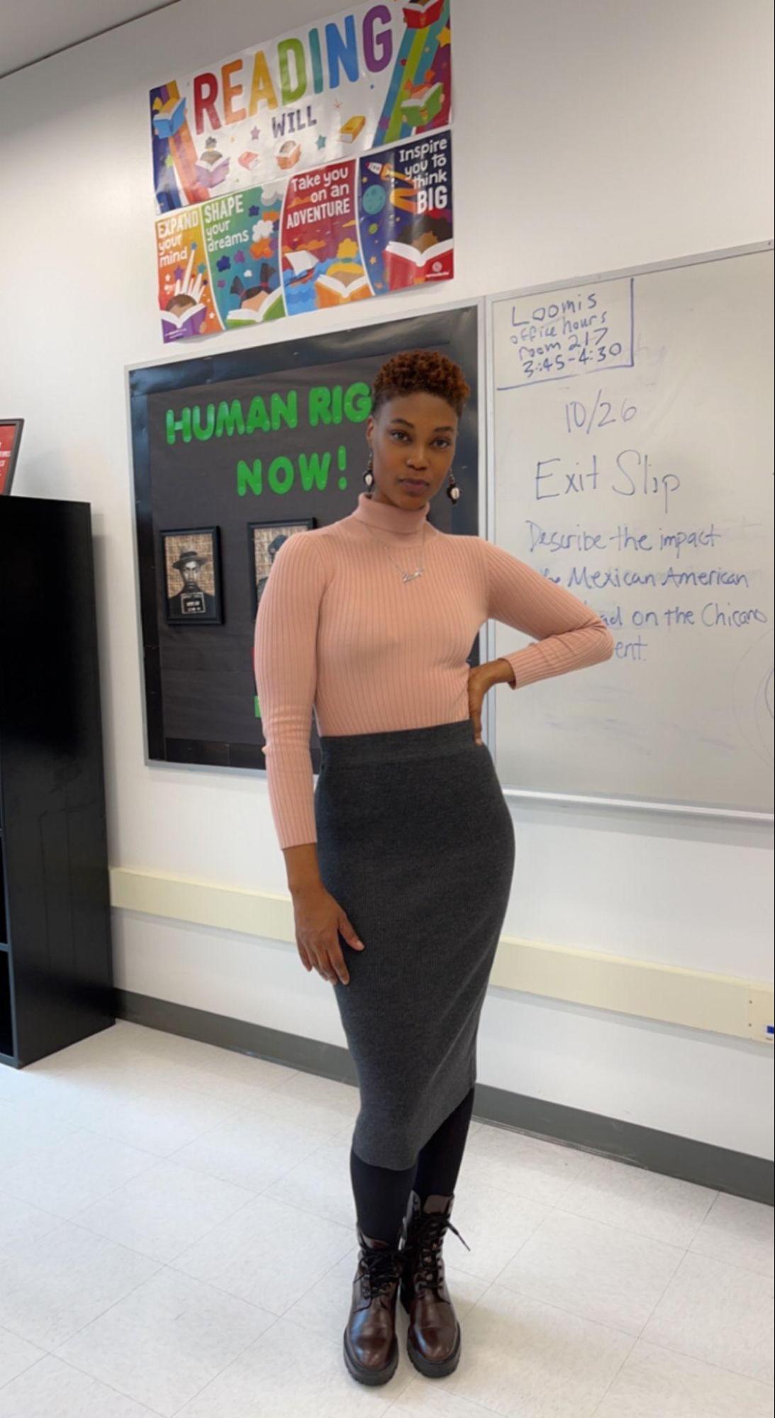 Ms. Emery is wearing a coral-pink turtleneck, long sleeved shirt. She wears a gray-ish long skirt with black leggings. She has her left hand on her hip as she poses for the photo.