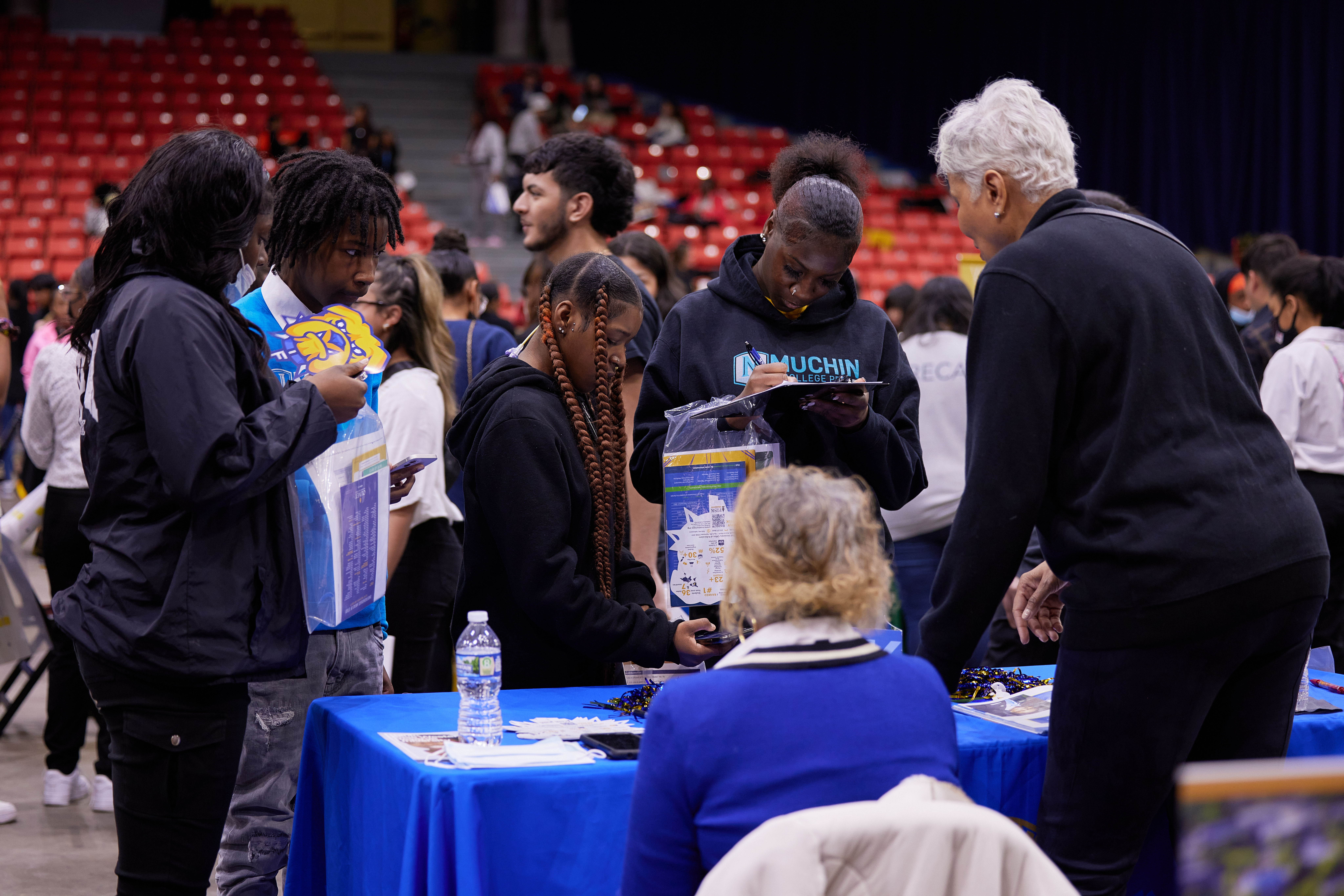 Photo shows a scene at the Noble Schools' College Fair at UIC's Credit One Arena. Two older Black women sit behind the Fisk University booth, which has a bright blue tablecloth. Several Black students from different Noble Schools stand in front of them, listening to them talk and looking at pamphlets and other materials on the table.