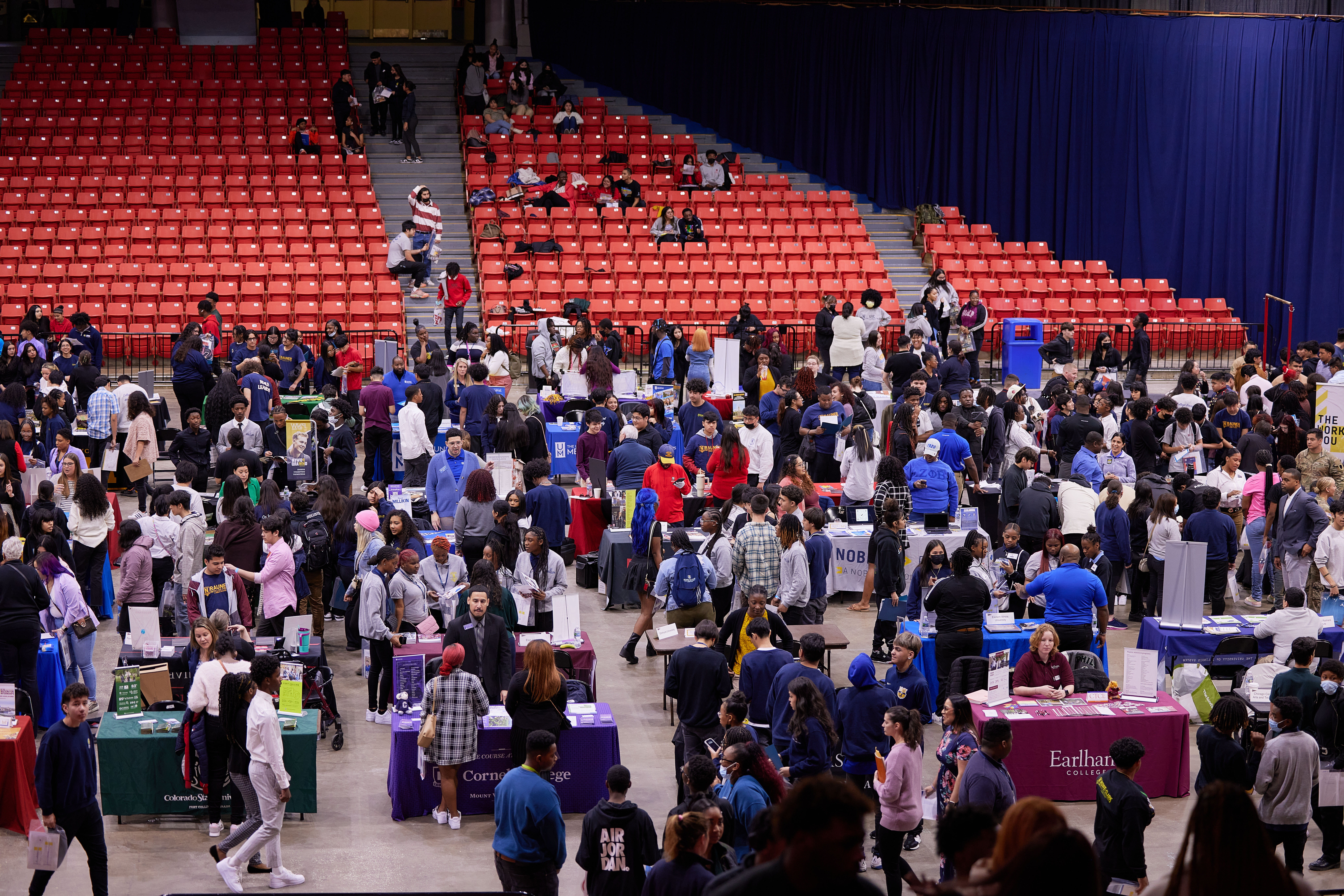 A wide shot of the Noble College Fair. On the top of the image, the orange seats are located. Some students are sitting down. Some students are sitting in the steps of the stairs. On the floor, there is crowded paths and booths filled with college recruiters and students.