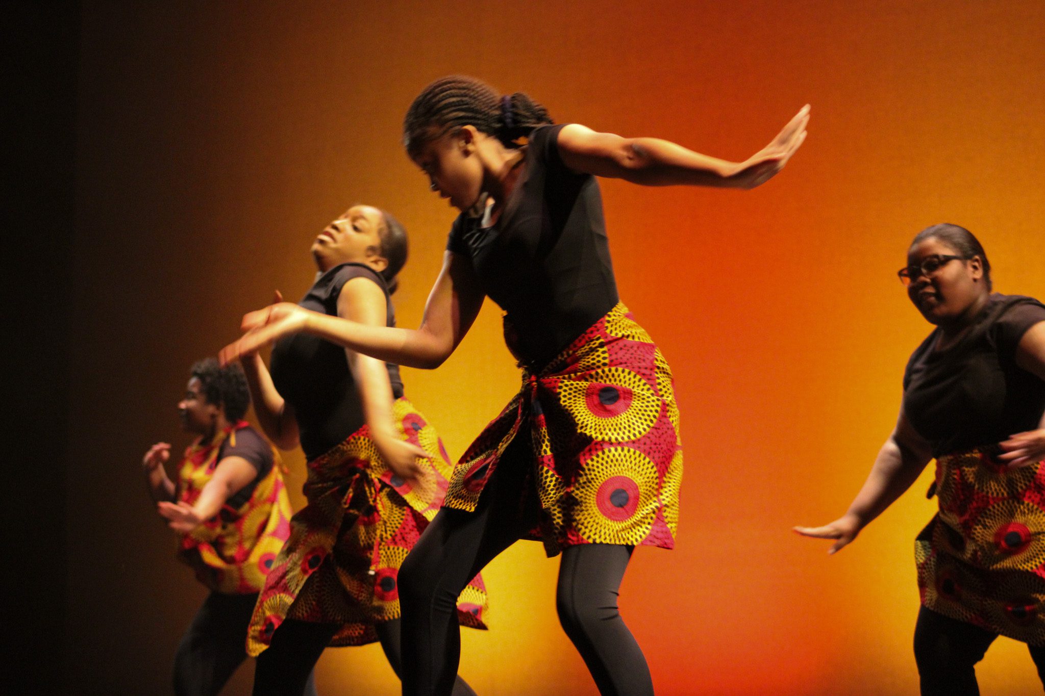 Four dancers are visible. The dancers are all wearing the yellow-orange circular patterned skirts. The dancers are all doing their dance moves as part of the routine. The background transitions into a black to orange shade.