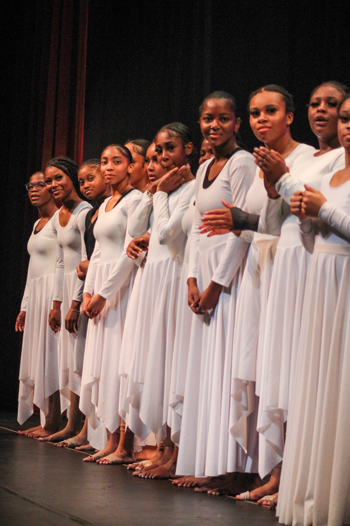 The dancers are all lined up in their dance uniform. They are all wearing a white ballet-like dresses. Some are clapping, while others are staring at the audience.