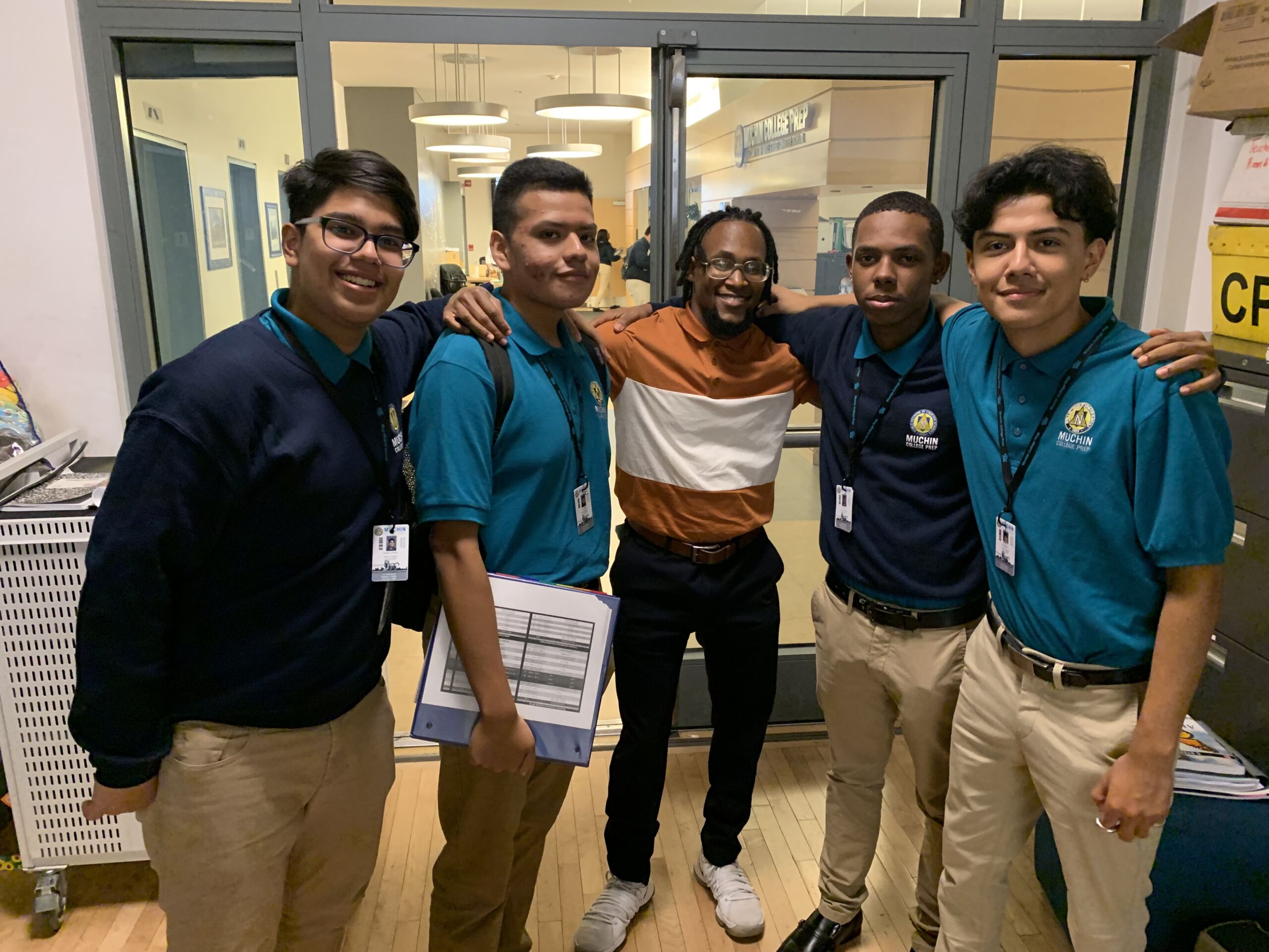 Emmanuel poses with his advisory class. There is two students posing next to him on the left and two students next to him on the right. The students are wearing the original Noble uniform with a navy blue sweater with khaki pants. Two students are wearing a teal button up school shirt. Emmanuel is wearing a half orange, half white stripped shirt.