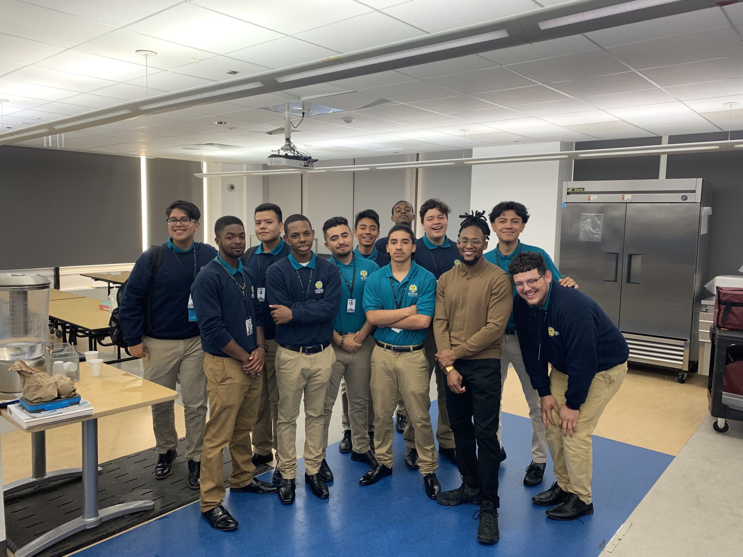 Emmanuel poses with his entire advisory. Emmanuel is wearing a beige sweater and has his hair wrapped up in a bun. The students are wearing navy blue sweaters and khaki pants. Some students are wearing teal button up school shirt.
