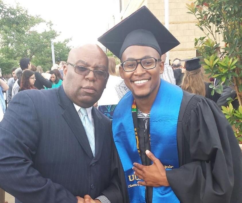 Emmanuel Jackson poses with his father after his graduation from Central Florida. His father, to his left, poses proudly with his son, shaking his hand in the photo. Emmanuel smiles at the camera with a thumbs up. He is wearing a blue graduation sash and black graduation cap and gown.