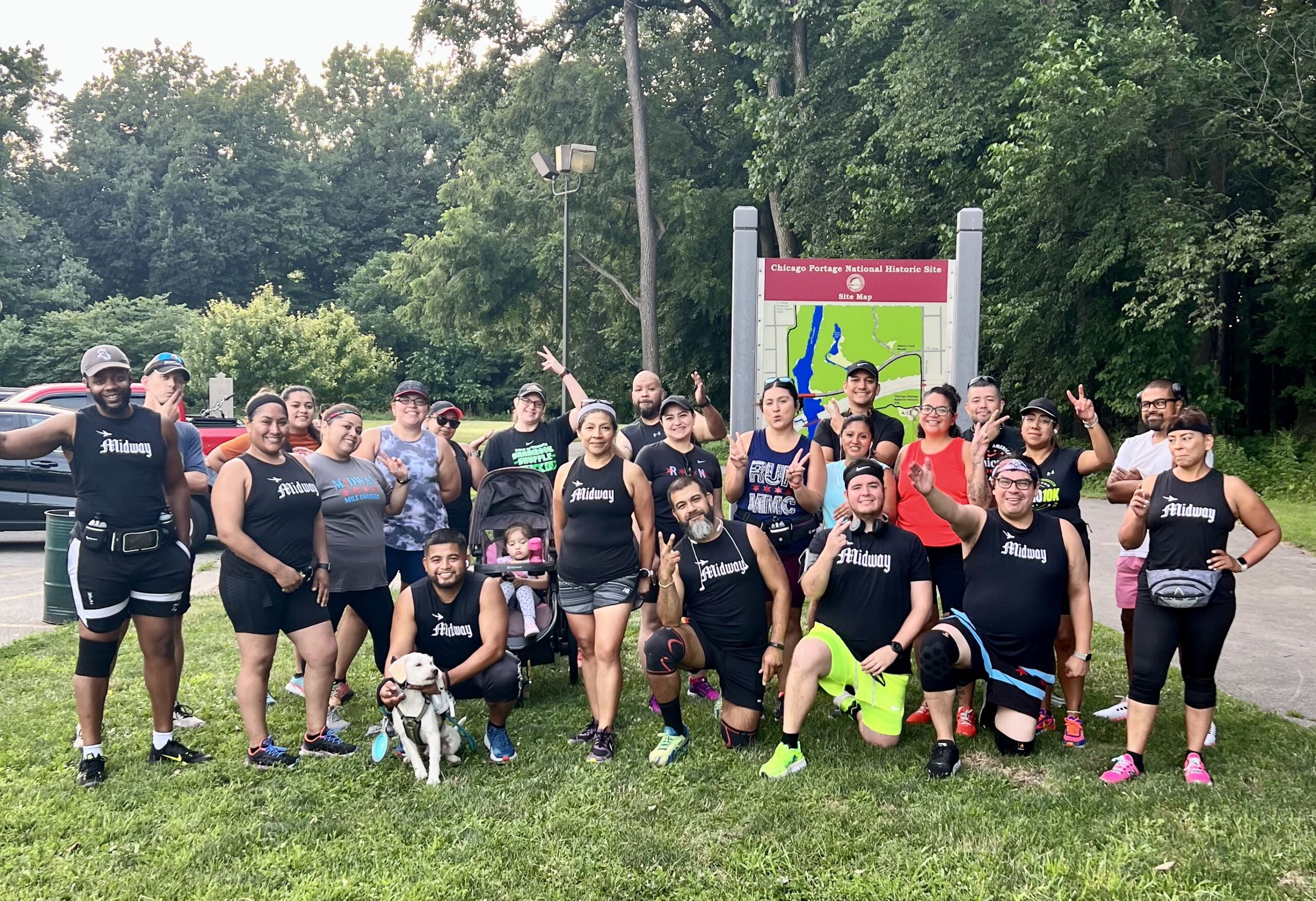 A group photo of the Midway Mile Chasers in front of the Chicago Portage National Historic Site where they had just gone for a long nature run. There are over 20 people smiling and holding up peace signs, almost all wearing matching black tank tops that say "Midway" on them in white text.