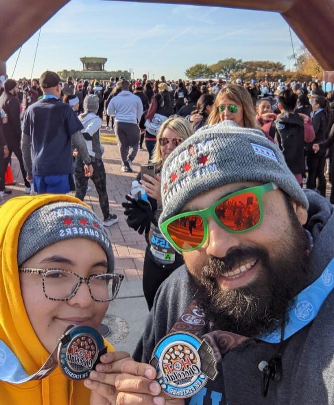 Photo shows a selfie of Mia and her dad holding medals for the Hot Chocolate 5K run in 2021. They are wearing matching gray beanies that say "Midway Mile Chasers" on them. Behind them is a crowd of other people at the race.