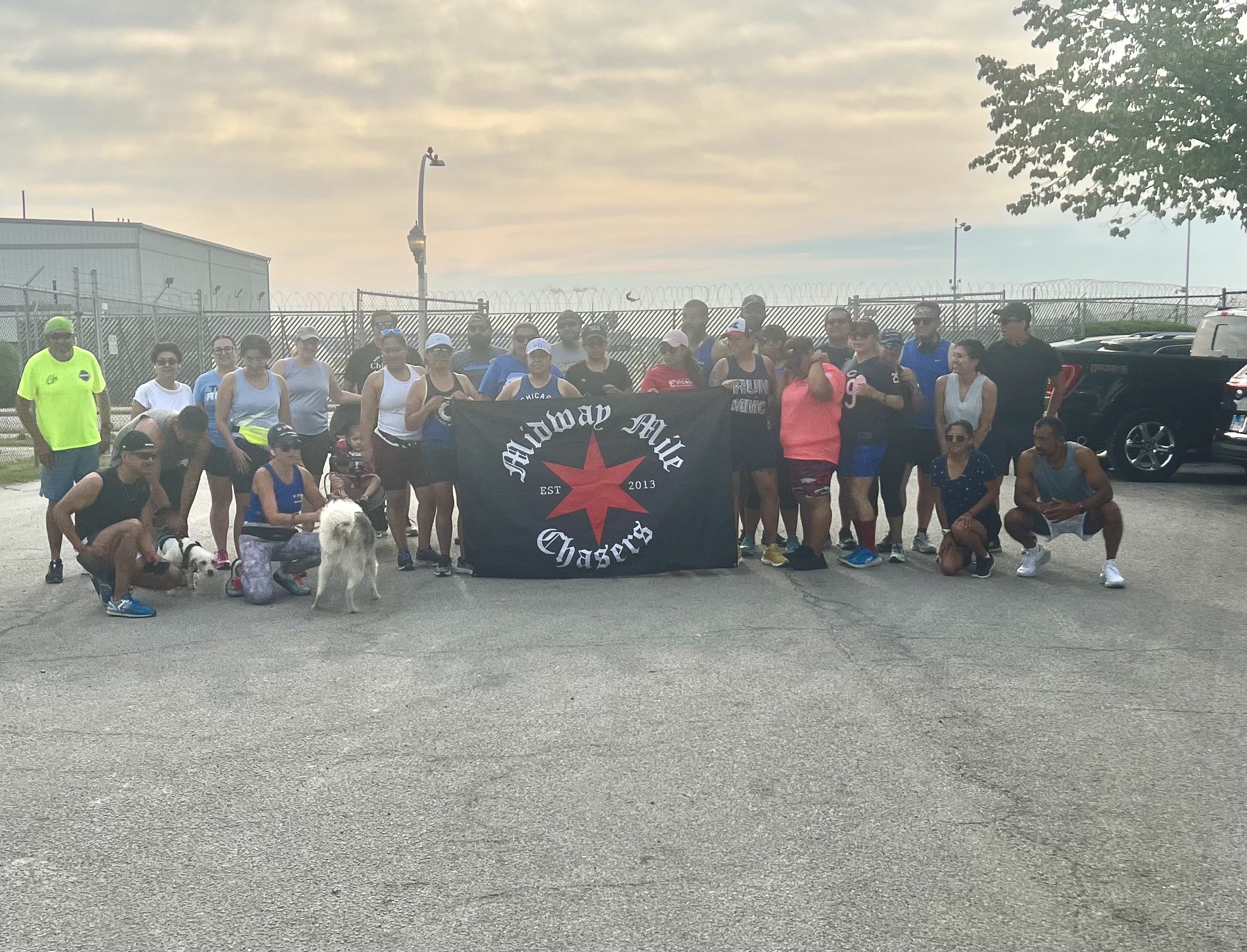 A group photo of the Midway Mile Chasers in front of the Midway International Airport. There are about 30 people posing together, holding a black flag in the middle. The flag has a big red Chicago star in the middle and has white text around it that says "Midway Mile Chasers, established 2013".