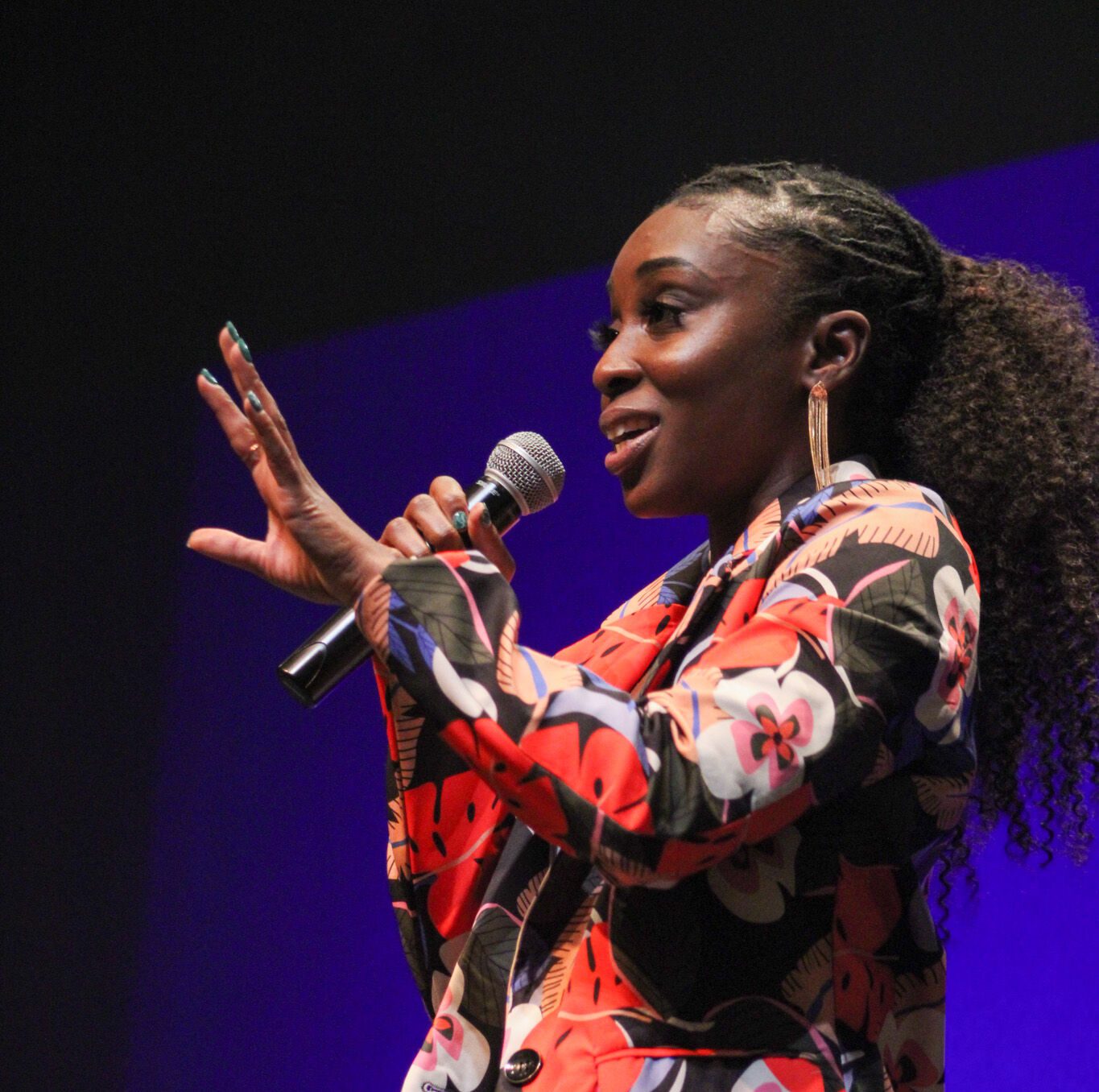 Ms. Lemon, dance instructor, is standing and facing towards the audience. She is wearing her hair braided to a puffy ponytail. She is wearing a orange-red floral patterned dress suit with blue-violet leaves. She is holding a microphone with her right hand.