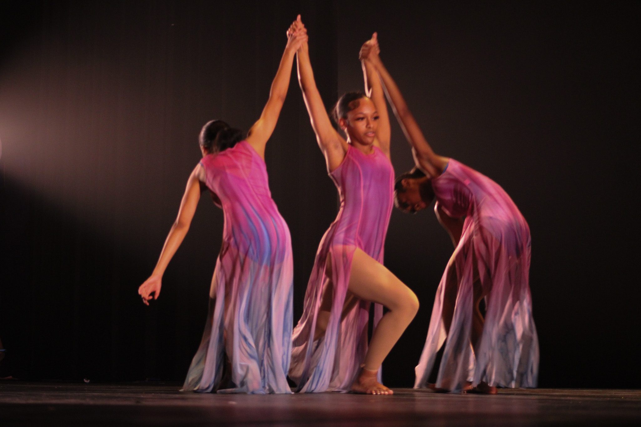 Three dancers are visible. Two of them are holding the third dancer as she moves forward. The dancers are wearing a gradient purple-white like dress. They have their hair pulled into a bun.