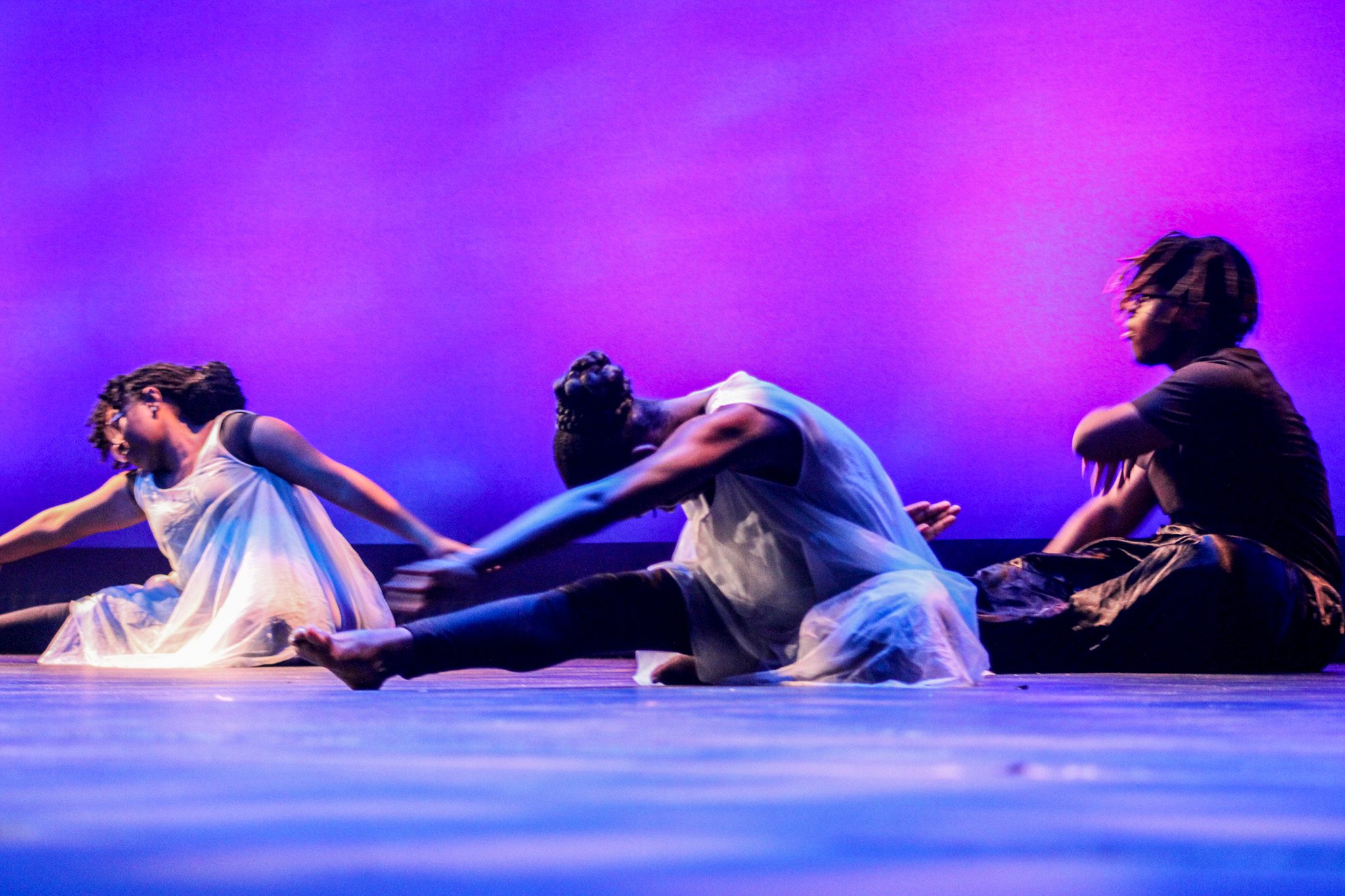 Three dancers are visible. The dancers are wearing a white, sheer-like dress, They are laying on the ground with their heads lowering to their body. They have their arms stretched out to touch their legs. The background is a gradient of purple and light purple.