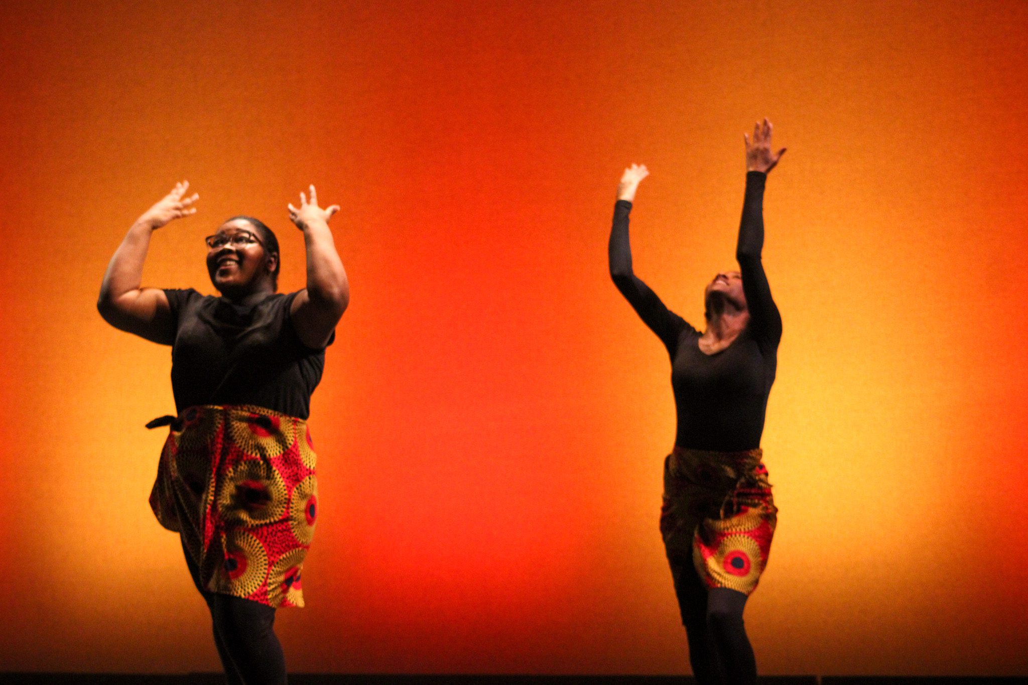 Two dancers from the Bengal Dance Collective. The dancer on the left is wearing a orange-yellow circular designed skirt with a small black t-shirt. She is smiling as her hands wave up to the sky, a part of the dance routine. The dancer on the right, wearing the same dance uniform. She has her arms up in the air and follows the dance routine. The background displays an yellow colored aura surrounding the two dancers. The other color transitioned into an orange color.