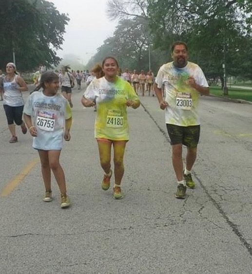 A photo of Mia and her parents running in a color run race when she was 10 years old. They are wearing matching white shirts, colored with the color run powder. They all have runner number tags on.