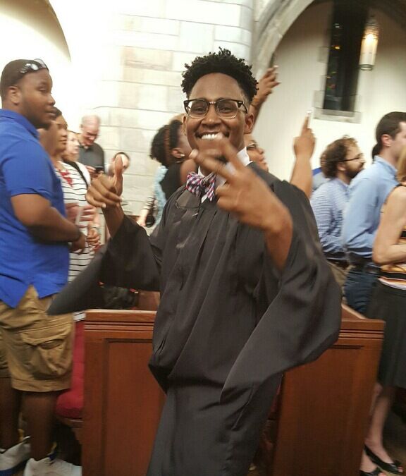 Emmanuel graduating from the University of Chicago. He is walking down the aisle as he poses, happily, with his hands up and smiling at the camera.