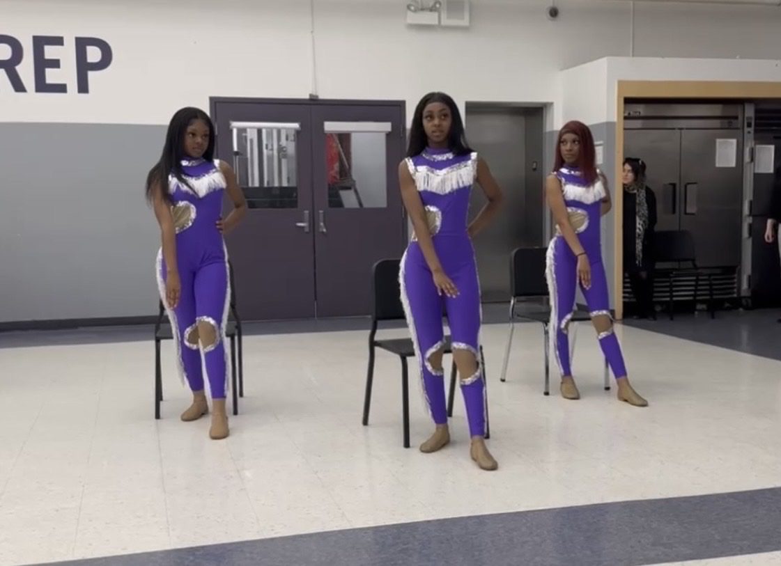 Three students are visible. They are wearing a one piece suit with purple and white colors. The dancers are in the middle of practicing their routine. The students are inside a building.