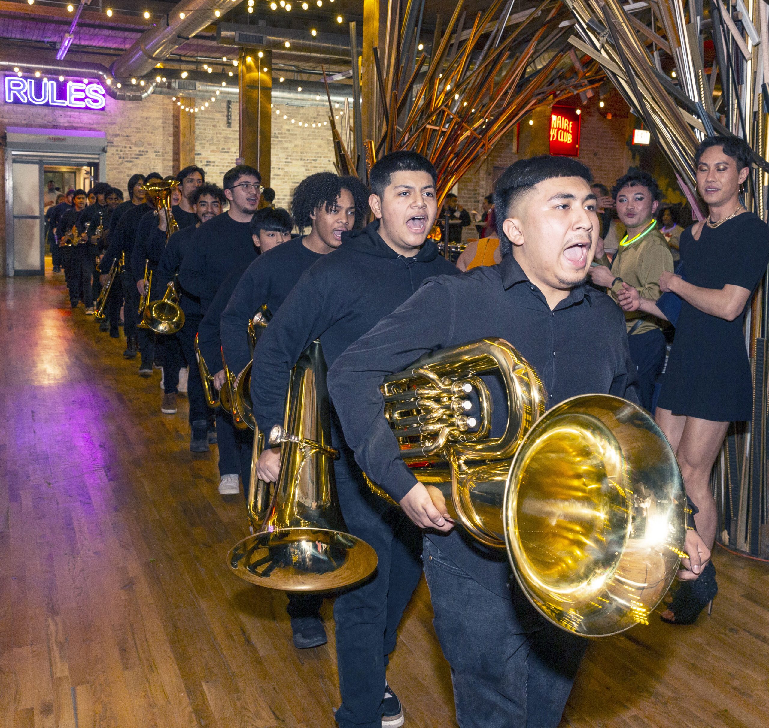 Tuba players, in a line, as they go about their performance. The first Tuba player, Fabian, is wearing a long black button up shirt and black dress pants. All the Tuba players behind are also wearing the same clothes. They are in a party hall with bright lights and golden decor.
