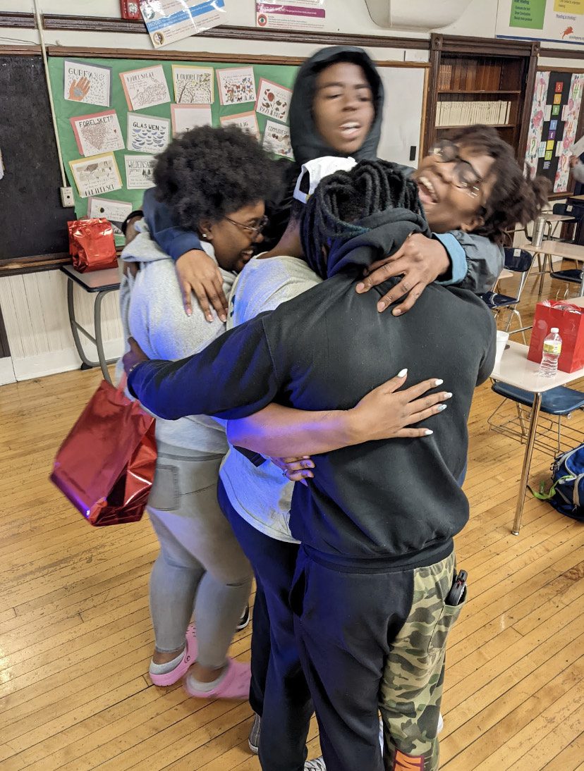 Photo shows five visible students hugging in a classroom. One student is wearing a hoodie on as he's hugging two of the students in the group. One student, who's face is not visible, is wearing his hoodie halfway. One student is wearing a white snap back hat but his face isn't visible. Two other students are both wearing glasses and another student is holding a red gift bag in her hand.