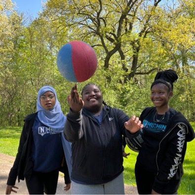 3 students are present in the image. One of the left is wearing a light blue head scarf and a Muchin school sweater. The student in the middle is wearing a black sweater and is spinning a basketball on his right finger. The basketball is half blue and half red. The student on the right has her hair in braids up in a bun. She is smiling at the student spinning the ball. She is wearing a black sweater with a Muchin shirt.