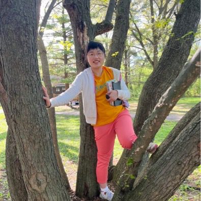 Student is posing in a tree. They are holding themselves on the tree with their right hand. They are wearing a white sweater with a yellow shirt and pink pants. They are also holding notebooks and books with their left hand. Their left leg is also placed in between tree branches.