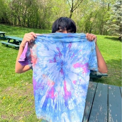 A student is holding a tie-dye t-shirt. The t-shirt is colored with blue, indigo, purple, and white colors. The student's face is not visible.