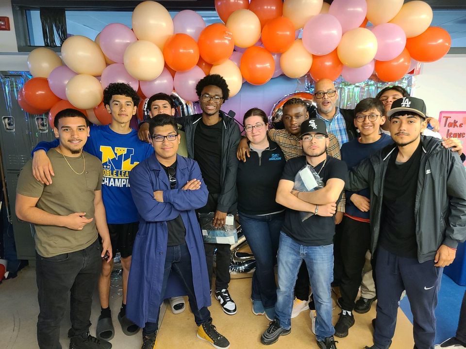 A group of male students pose together at Senior brunch. They are pose holding onto each other. Some students have their arms folded and surrounded by staff members. Behind the students is orange and light pink balloons.